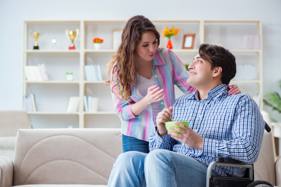 A man in a plaid shirt sitting in a wheelchair at home, chatting with a support worker