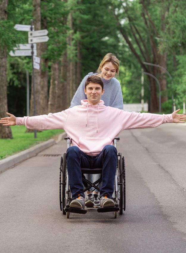 A smiling young man in a pink hoodie in a wheelchair with arms outstretched, pushed by a support worker along a forest road